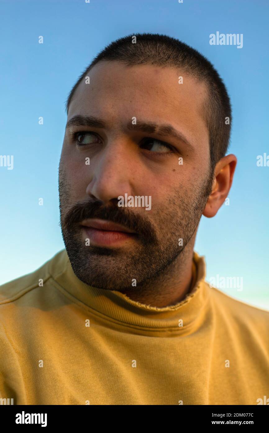Portrait of young man with moustache looking away in front of blue sky wearing yellow jumper Stock Photo