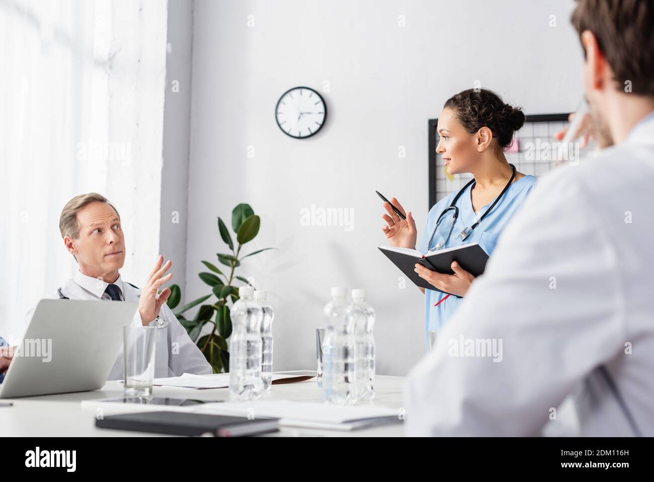 African american nurse with notebook and pen looking at doctor with raised hand near colleague on blurred foreground Stock Photo