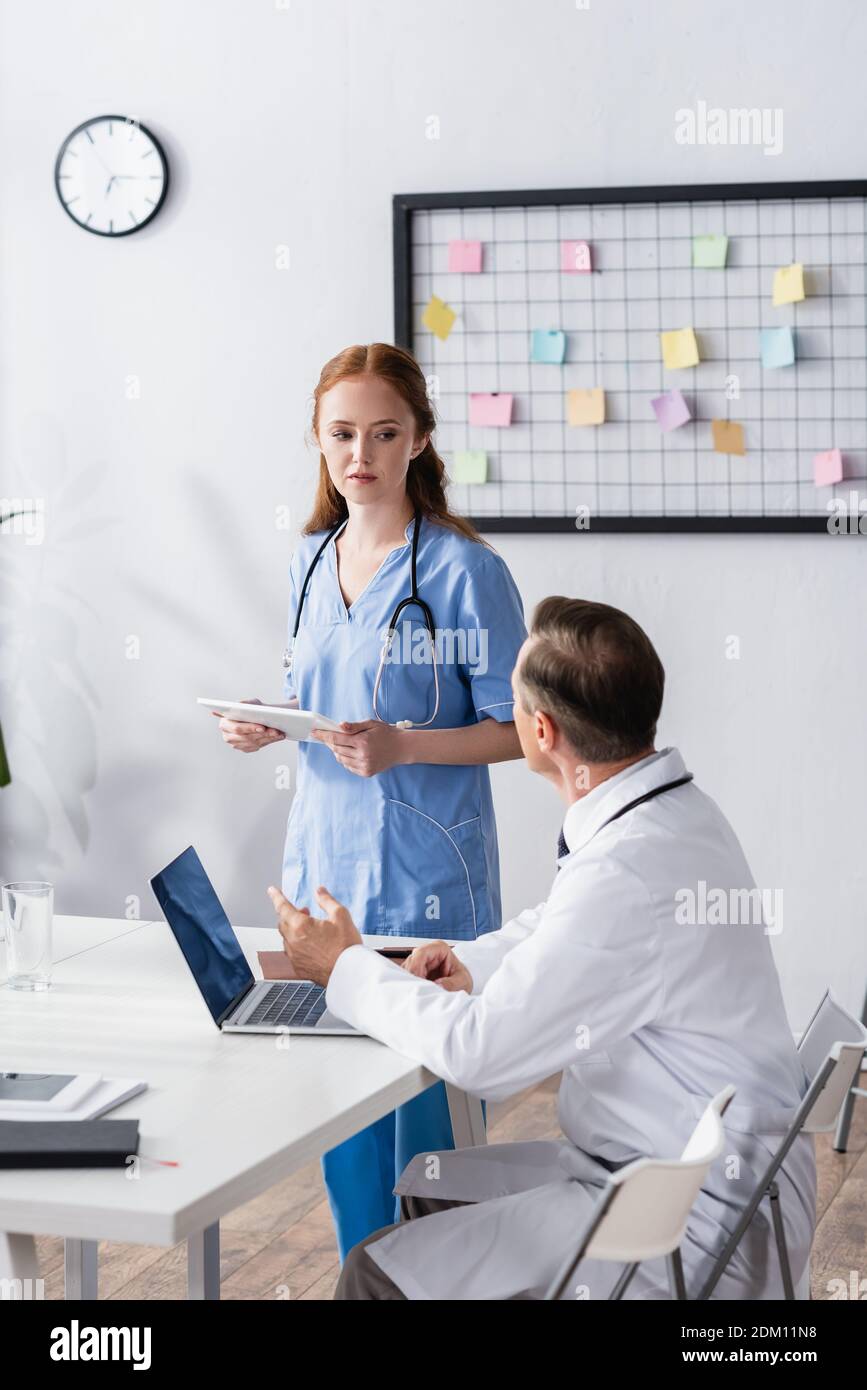 Nurse with digital tablet looking at doctor near laptop and notebook on blurred foreground Stock Photo