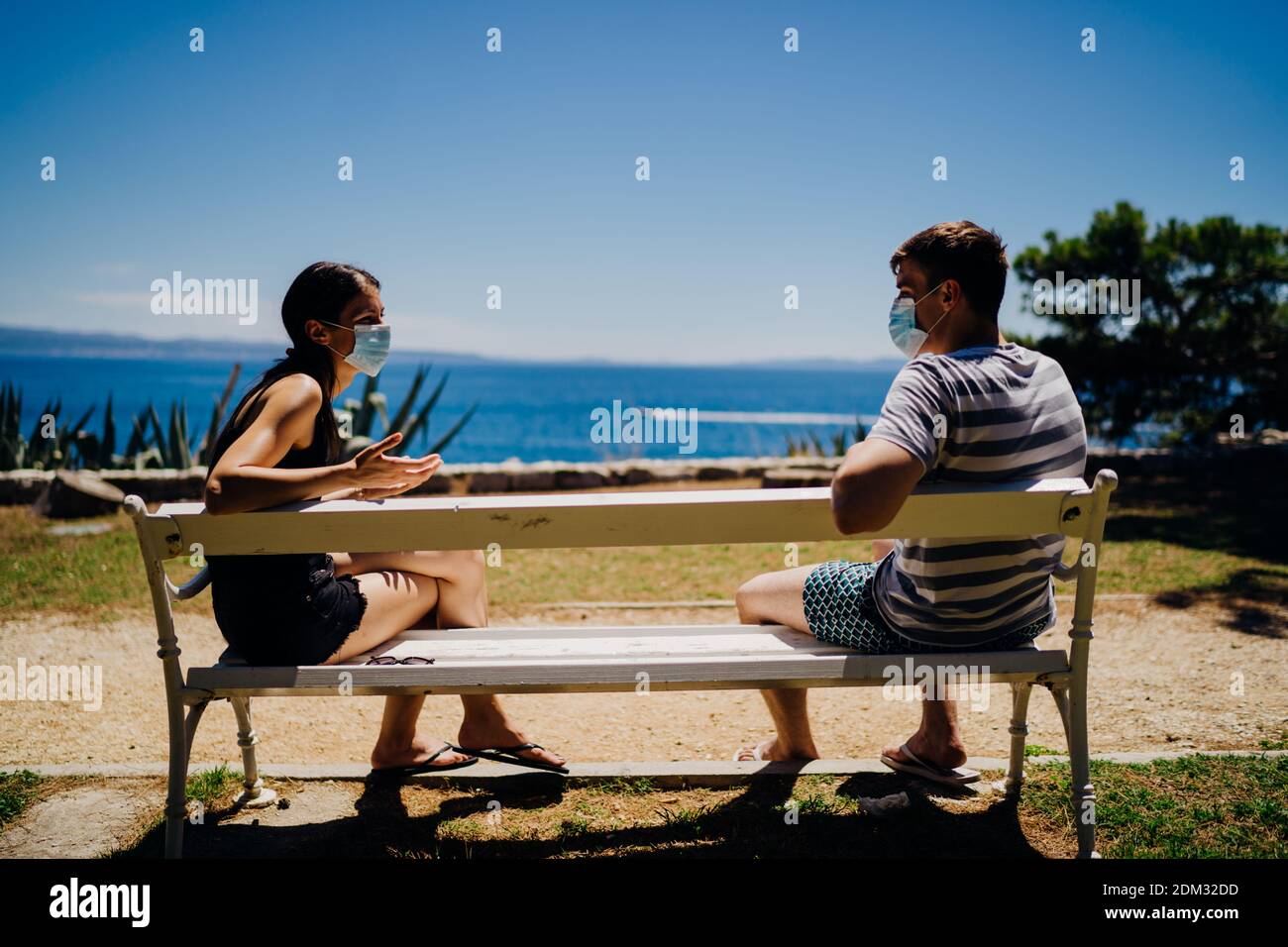 Young couple having an outdoor date on a bench in a park.Social distancing dating.Friends talking, wearing protective face masks.Coronavirus pandemic Stock Photo