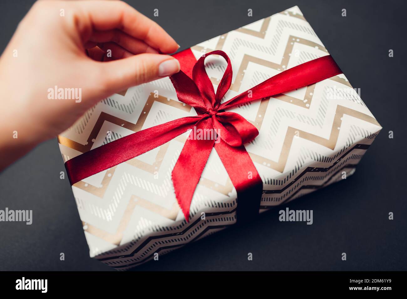 Woman opens Christmas and New year gift box wrapped in paper and decorated with red ribbon bow on black background. WInter holiday present Stock Photo