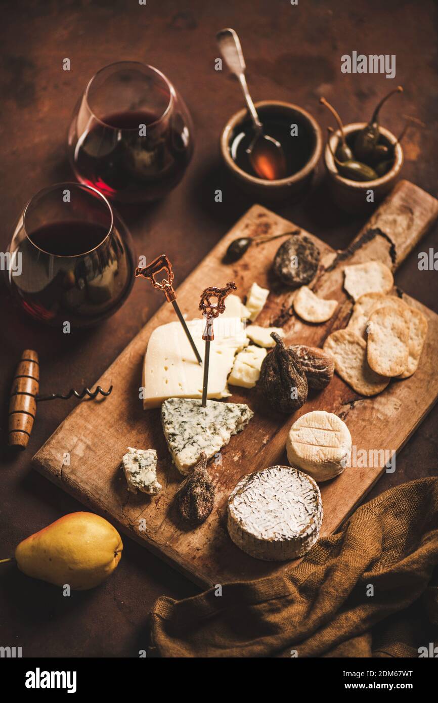 Assortment of snacks and appetizers for red wine concept. Various cheeses, crackers, dry and fresh fruit and glasses of red wine on wooden rustic board over rusty brown background Stock Photo
