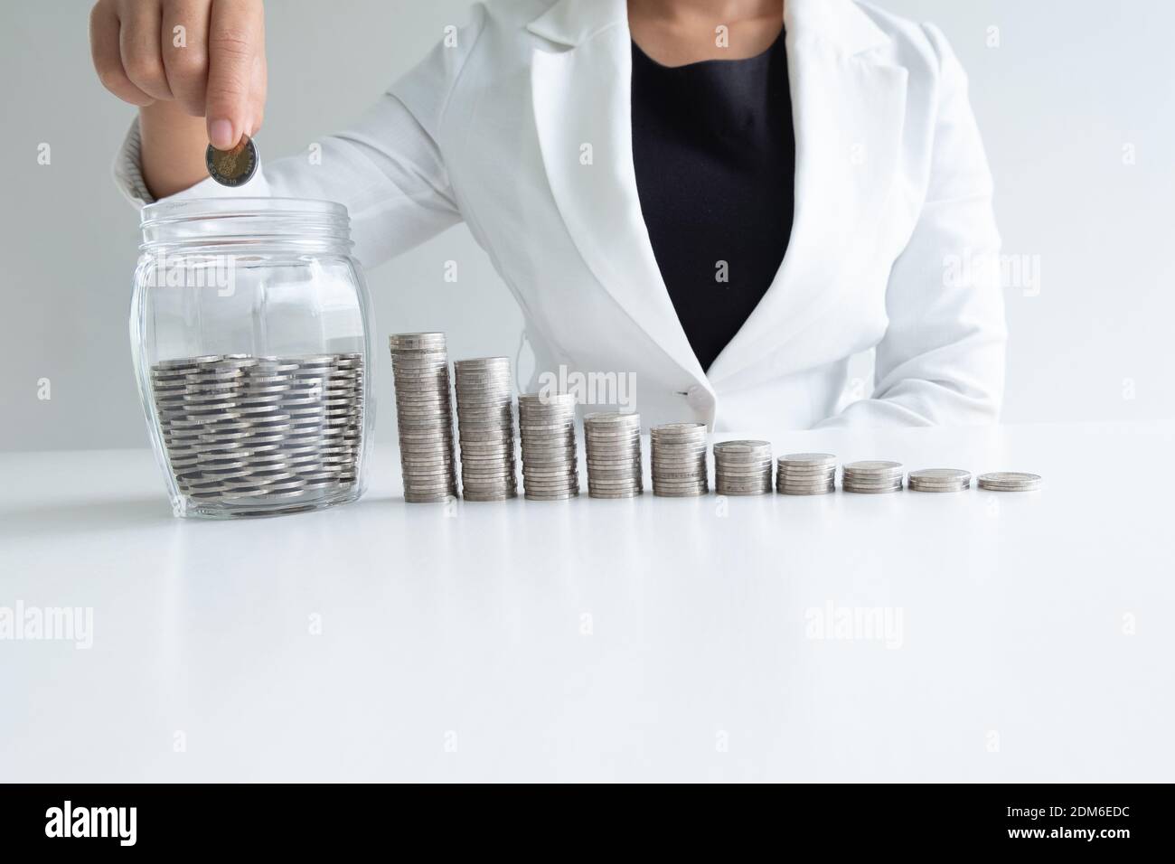 Midsection Of Businesswoman Putting Coin In Jar At Desk Against White Background Stock Photo