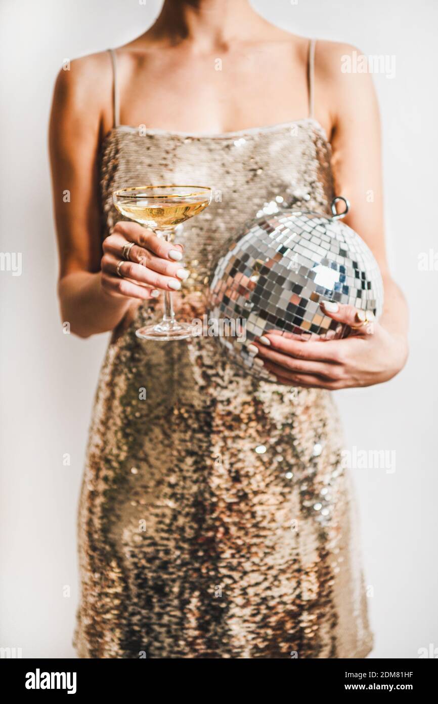 Young woman figure in festive glittering cocktail golden mini dress holding glass of champagne and discoball over white wall background. New Year holiday party concept Stock Photo