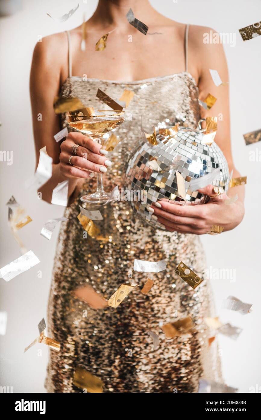 Young woman figure in festive glittering cocktail golden mini dress holding glass of champagne and discoball in cloud of golden confetti, white background. New Year holiday party concept Stock Photo
