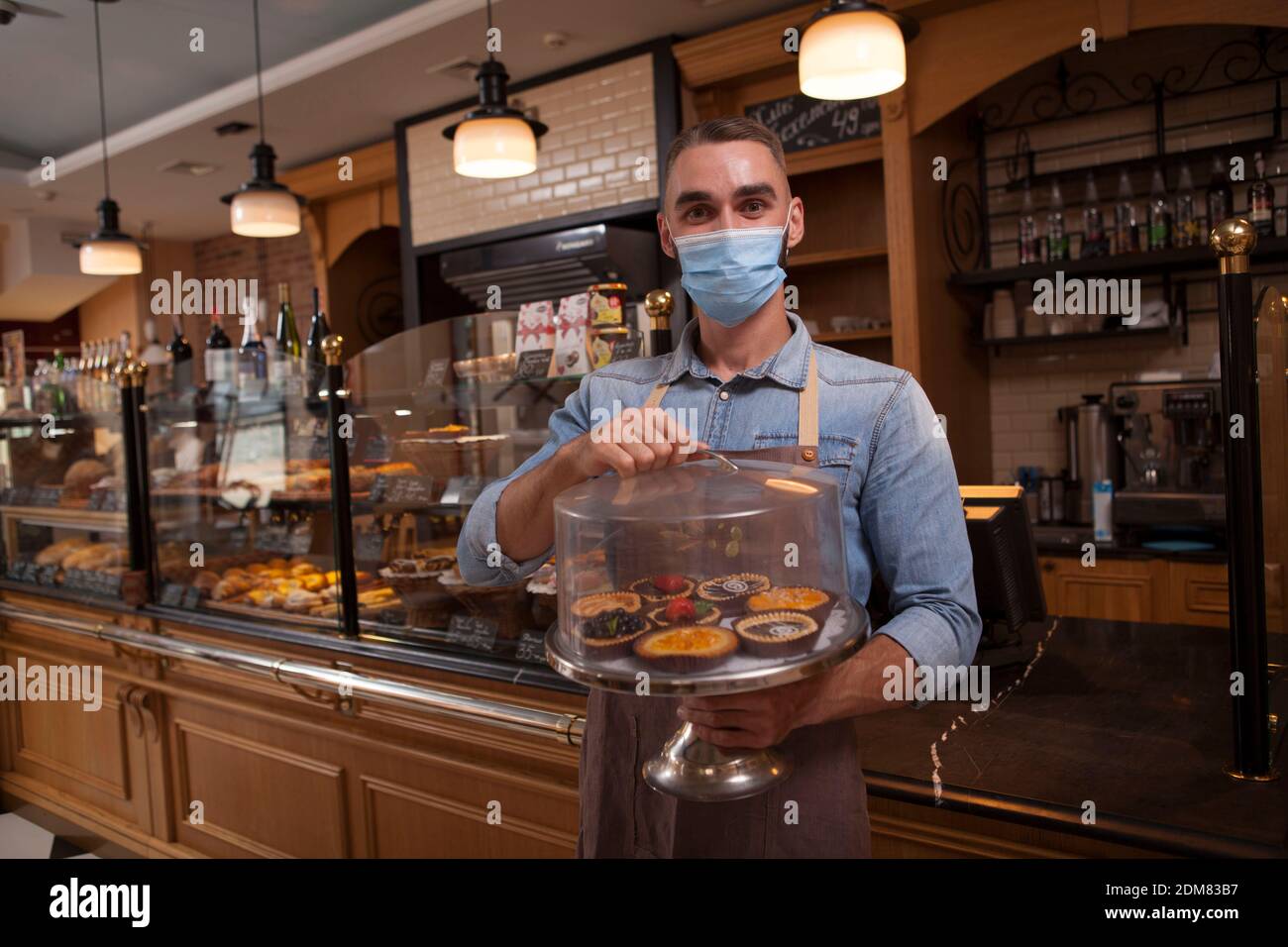 Cheerful male baker wearing medical face mask, working at hic coffee shop selling delicious