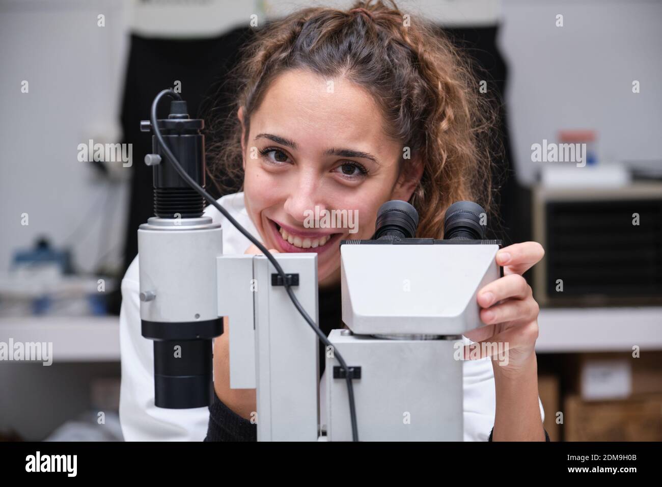 Young female scientist looking at camera and smiling next to a microscope in a laboratory. Laboratory research concept. Stock Photo