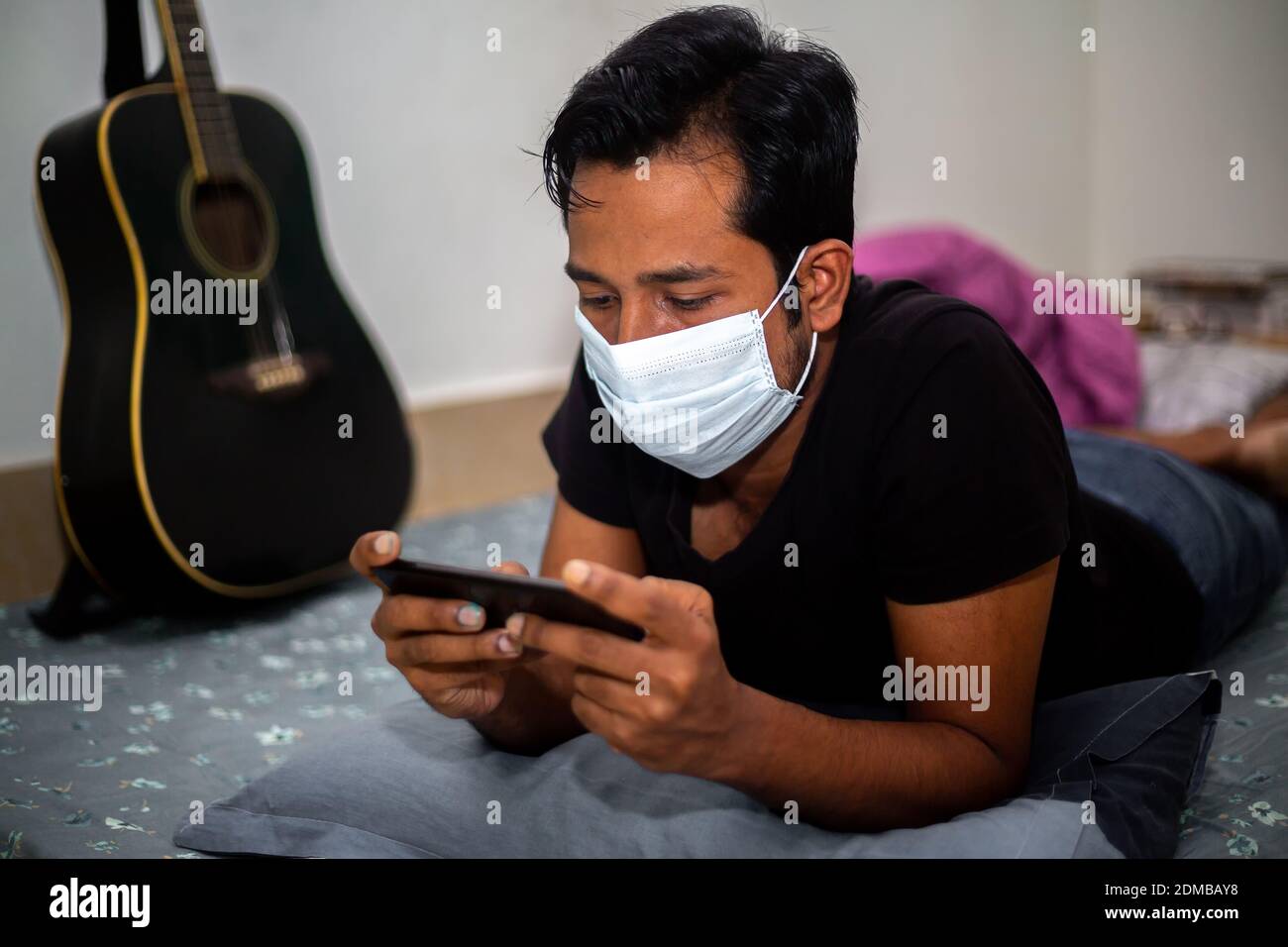 A surgical mask-wearing young man was playing mobile games on his own home due to coronavirus home-quarantine at Dhaka. Stock Photo