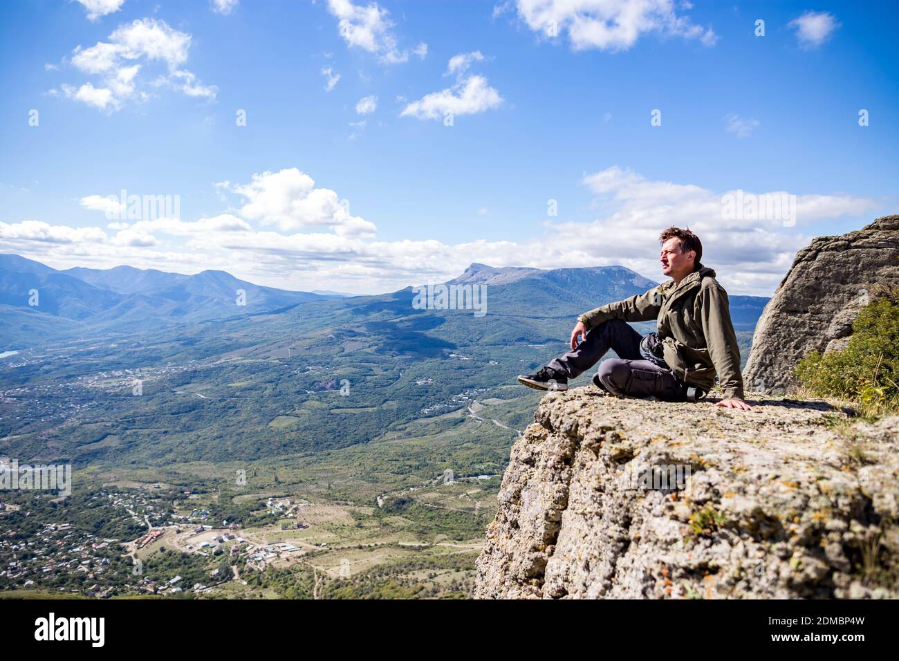 Male Hiker Sitting On Cliff Against Blue Sky Stock Photo