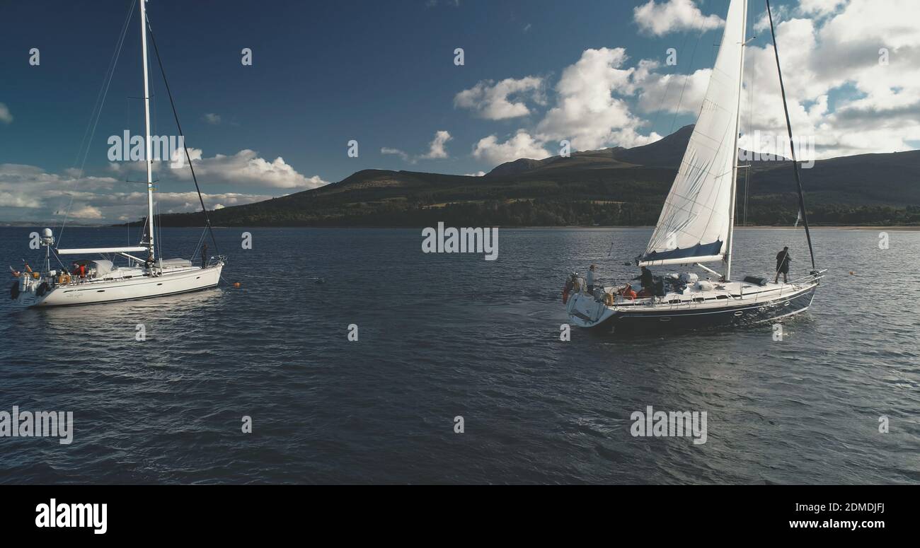 Closeup of motor boat sails at yachts on ocean bay aerial. Sunlight over serene seascape with water transport. Green mountain shore of Brodick harbor, Arran island, Scotland. Cinematic soft light Stock Photo