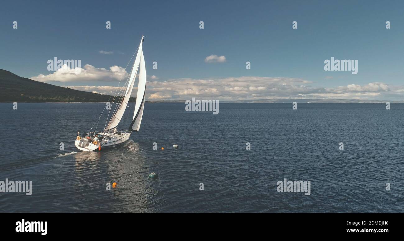 Yacht sail cruise aerial. Sailboat reflect at ocean water. Mountain coast of Brodick Gulf, Arran island, Scotland, Europe. Fluffy clouds float at windy summer day. Cinematic drone shot Stock Photo