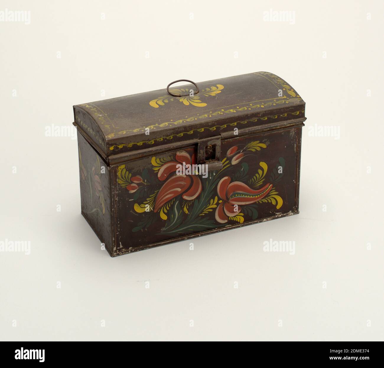 Box, Tôle (painted tin), Rectangular box with hinged cover domed from front to back. Ring bail handle on top of cover and hasp fastener, with staple broken off. Box is painted black with floral decoration on front and ends of vermillion fruit-like flowers, with white and alizarin markings, and with serrated green leaves and leaf-fronds. Cover is decorated on front and ends with a border of lapped and recumbent 'S's in yellow and similar band with dots frames the top. Spiral arrangement of leaf-fronds in yellow around handle., New York, USA, early 19th century, metalwork, Decorative Arts, Box Stock Photo