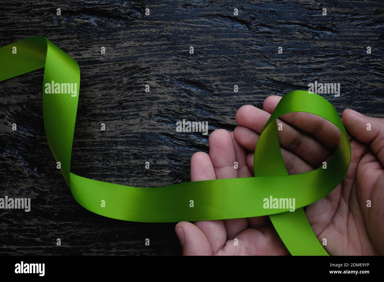 Top view of hands holding lime green color ribbon on dark background. Non-hodgkin lymphoma cancer, lyme disease, muscular dystrophy and postpartum Stock Photo