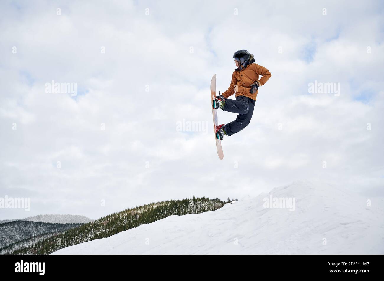 Guy snowboarder in winter jacket and helmet jumping in the air. Adorable kid making jump with snowboard while sliding down snowy hill in winter mountains. Concept of winter sport activities. Stock Photo