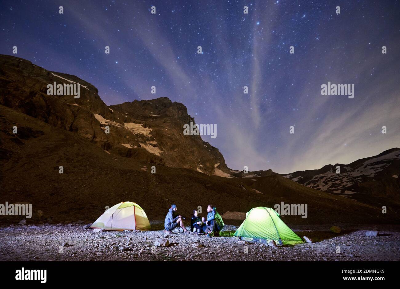 Night Starry Sky Over Mountains With Group Of Hikers Near Illuminated Camp Tents Tourists Sitting Under