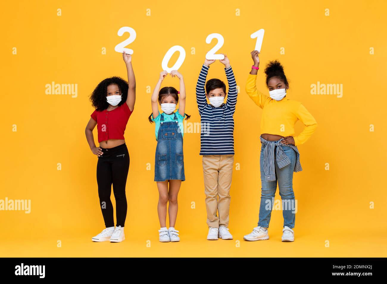 Cute mixed race children wearing medical face masks holding 2021 numbers isolated on yellow background, new year in the time of pandemic concepts Stock Photo