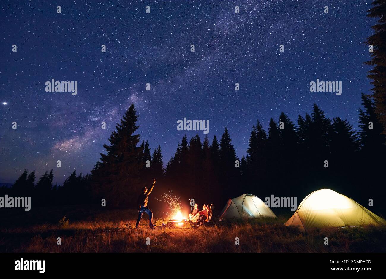 Man showing his friends Milky Way over tent city. Guys are sitting by the campfire on the background of the spruce forest and enjoying the evening starry sky. Outdoor recreation, touristm concept Stock Photo
