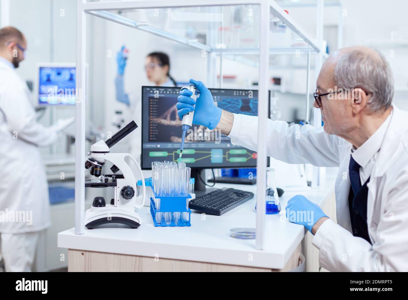 Experienced scientist working in busy laboratory at his workplace holding dropper dispense. Senior professional chemist using pippete with blue solution for microbiology tests. Stock Photo
