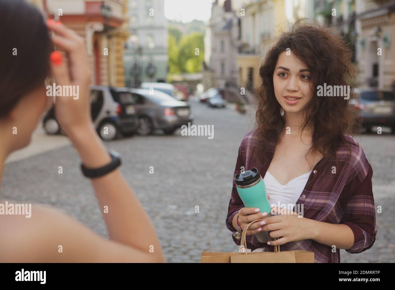 Two young women talking, enjoying warm day, wandering in the city. Lovely woman smiling at her friend while talking on city streets Stock Photo