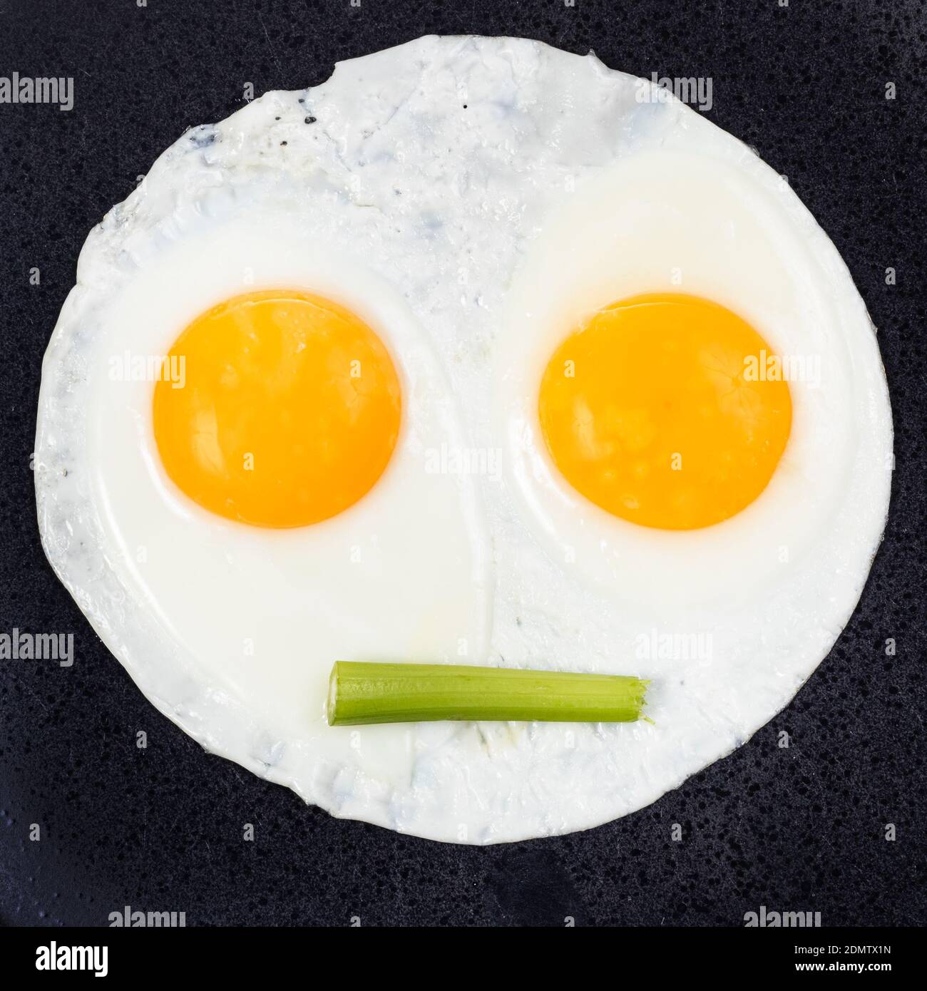 fried eggs and piece of celery on black plate close up. Fried eggs like face with straigh closed mouth Stock Photo