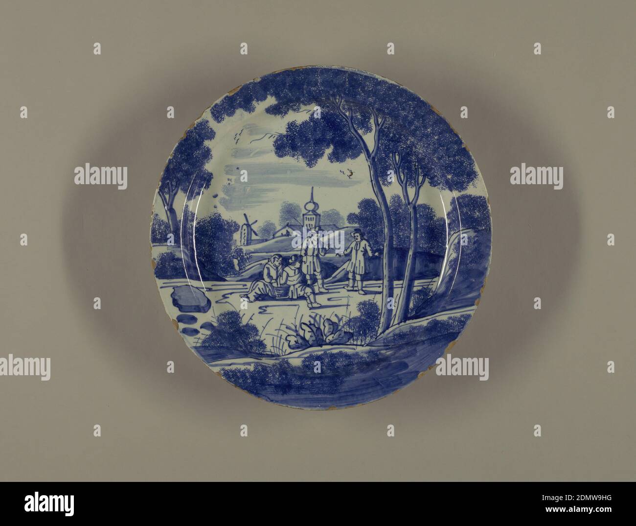 Plate, Tin-glazed earthenware, Circular plate with deep cavetto, standing on short foot rim; painted in underglaze blue on white with overall scene of 4 figures in landscape (two seated, two standing), 2 large trees on right, a windmill and onion-domed tower background, spongework leaves., Delft, Netherlands, ca. 1720–40, ceramics, Decorative Arts, Plate Stock Photo