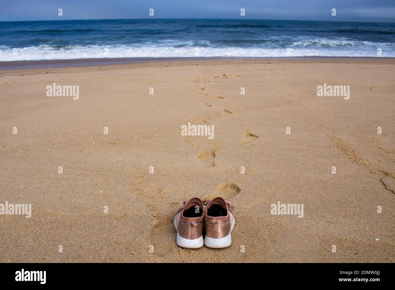 Pink canvas shoes resting on the sand facing the ocean at empty beach with footprints and waves in the background Stock Photo