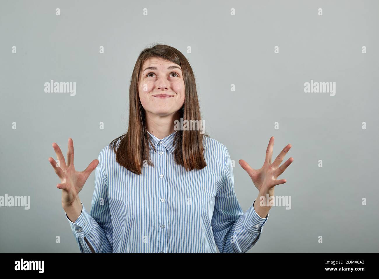Palms up, place your product here, raising arms wide spread is catching something. Young attractive woman, dressed blue shirt with brown eyes, brunette hair, grey background Stock Photo