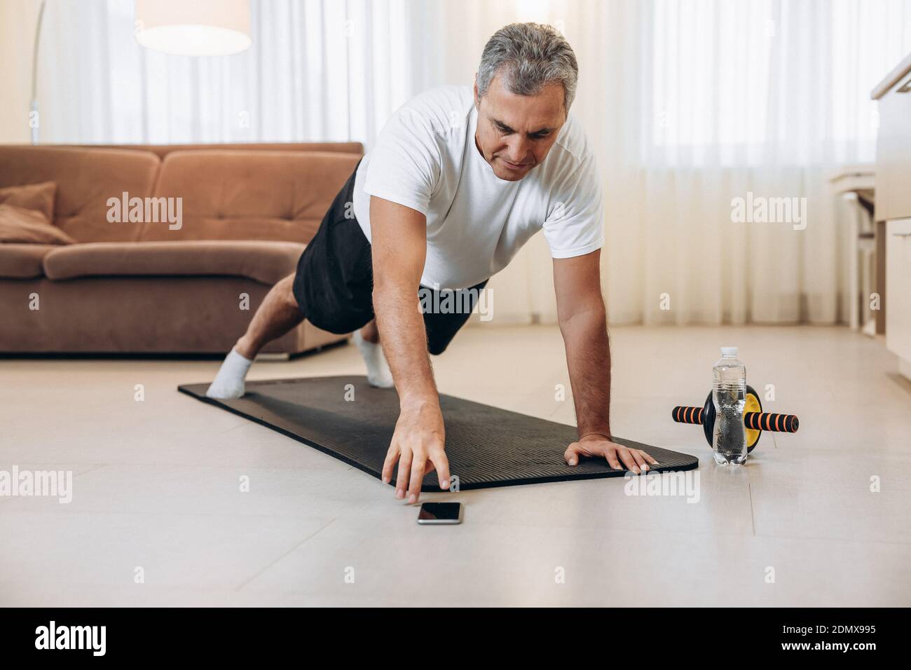 Elderly man reaches for phone to change music during excercise. Workout on yoga mat at home. Old man stands in plank position. Morning exercises with Stock Photo Alamy