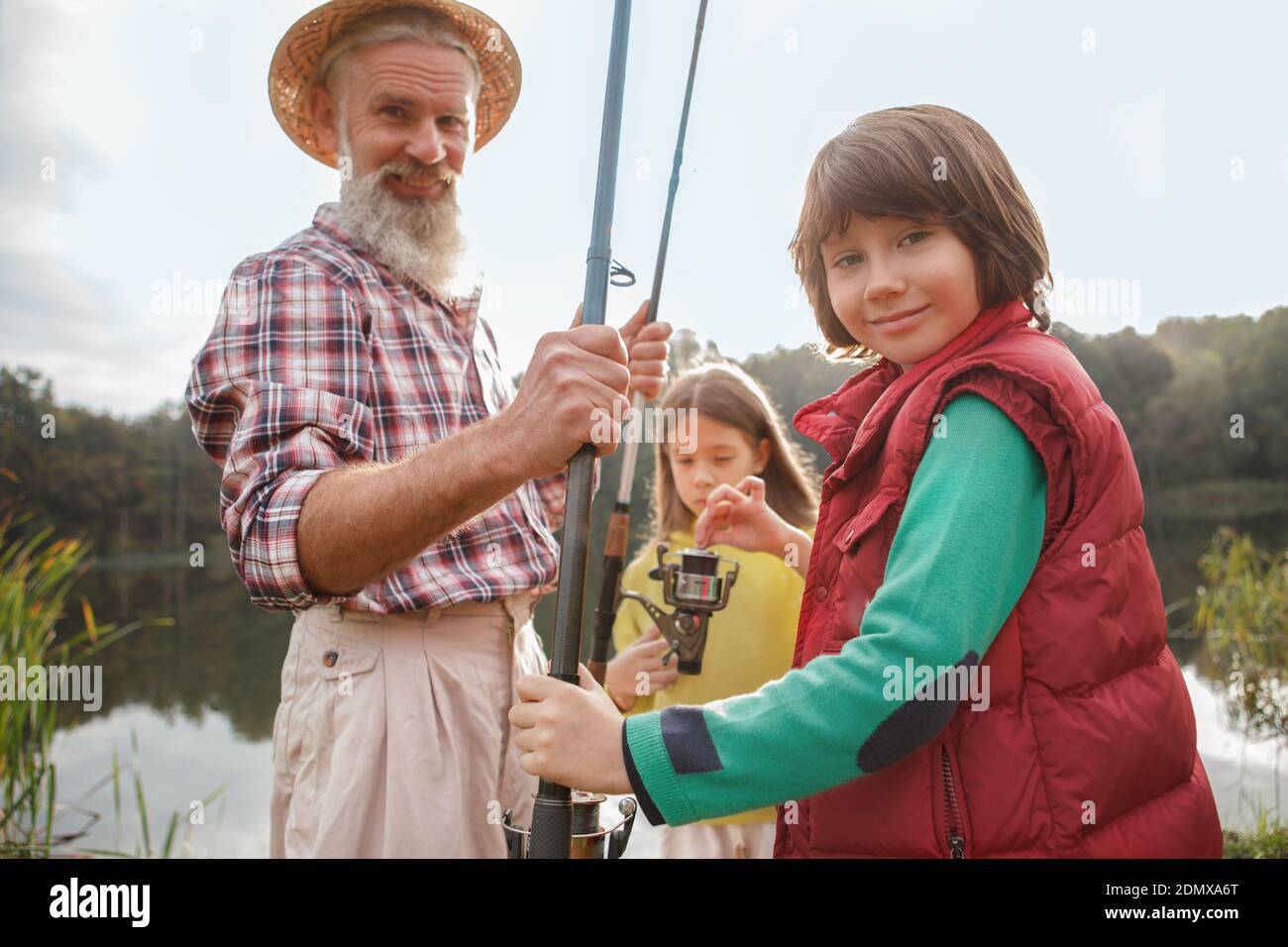 Cute happy young boy smiling to the camera, enjoying fishing with his grandfather and sister on the lake Stock Photo