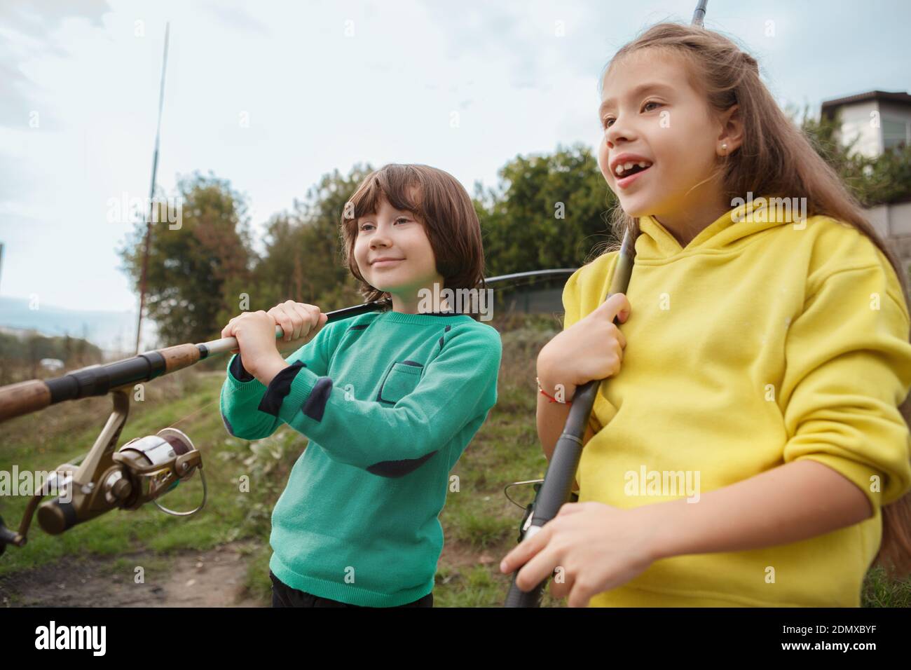 Adorable happy children holding fishing rods looking away at the lake cheerfully Stock Photo