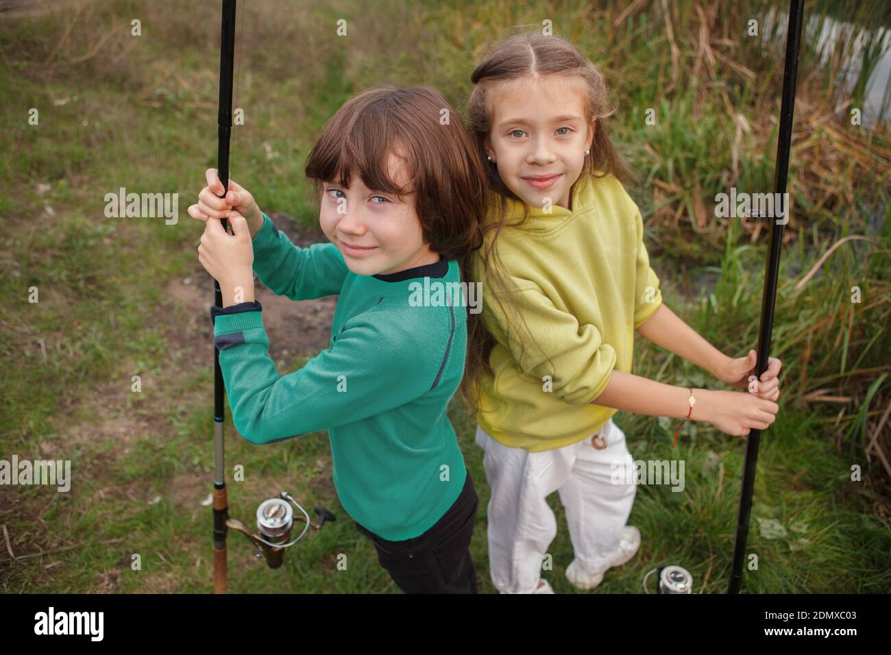 Top view shot of cheerful young boy and girl smiling to the camera, standing back to back holding fishing rods outdoors Stock Photo