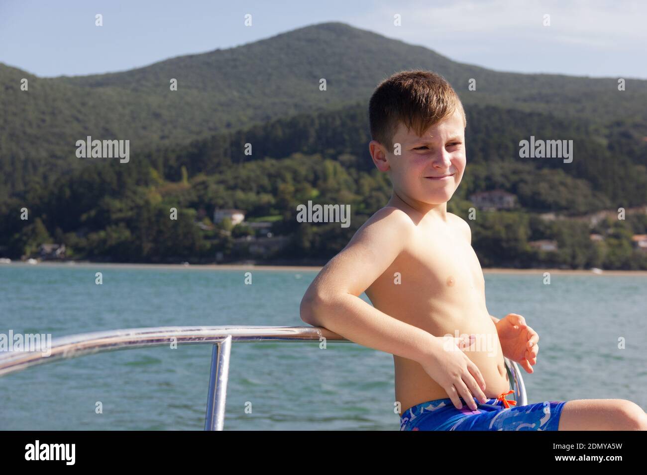 White skinny boy on blue swimming trunks leans on boat railing by the sea with strong sun on chest. Child sunbathing sitting on small vessel prow Stock Photo