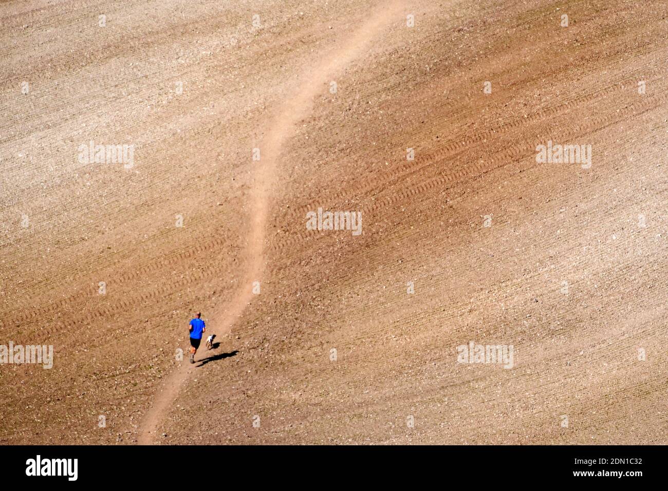 Man and his dog running up a hill on winding path across a vast field Stock Photo