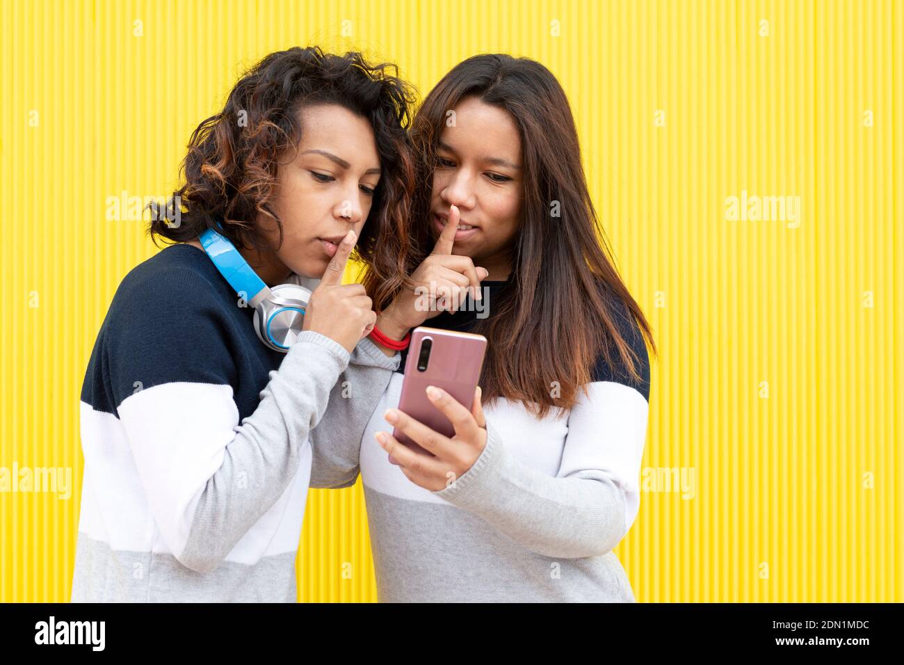 Portrait of two brown girls on a yellow background. Both are making the gesture of asking for silence while looking at a smart phone. Space for text. Stock Photo