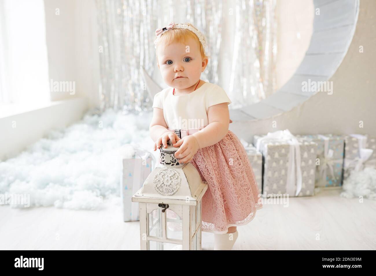 Cute baby girl in a pink dress and with a pink bow is standing in light ...