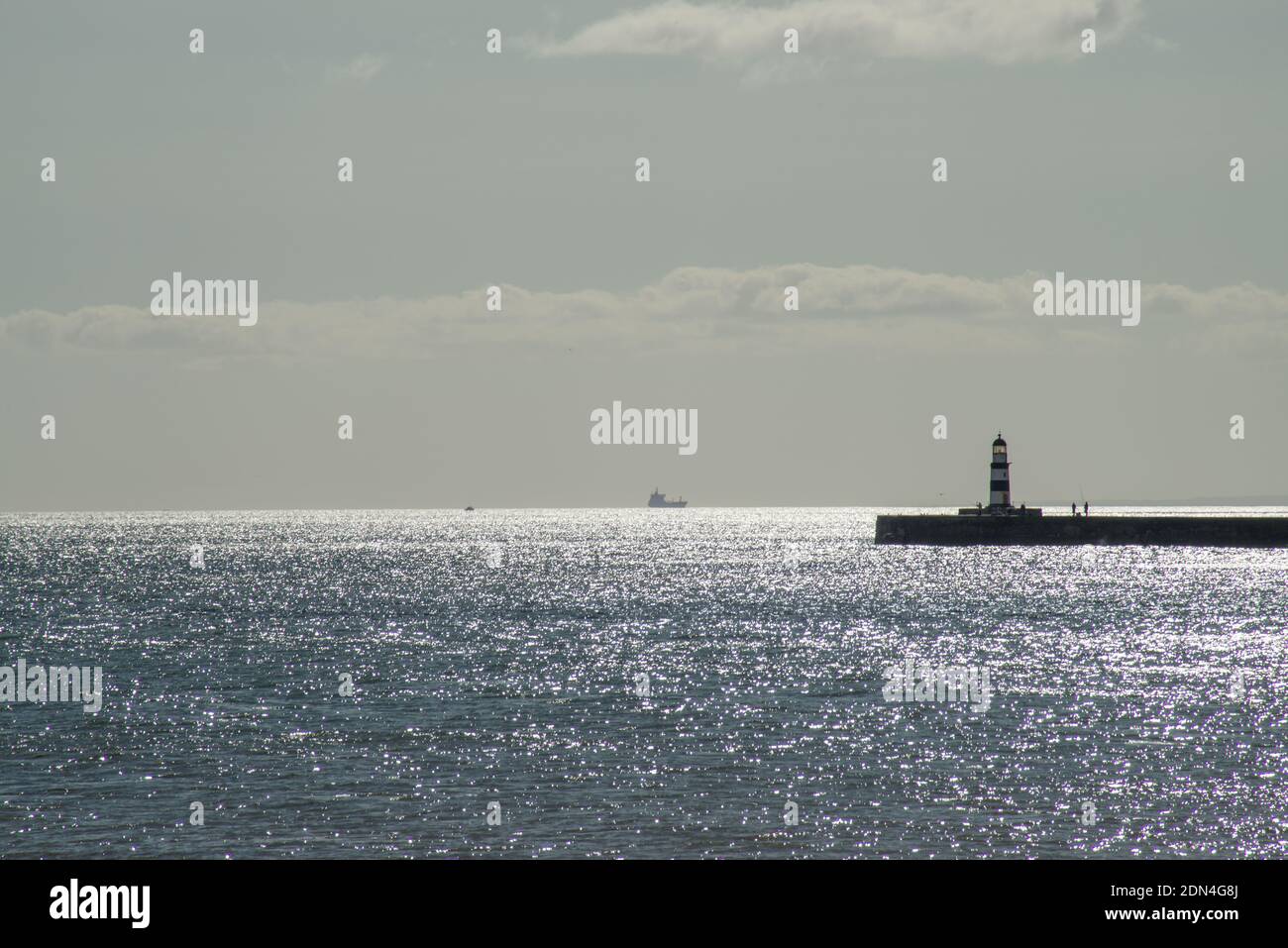 Image of late afternoon, with sunlight glinting on a calm sea at the entrance to Seaham Harbour with a ship out at sea and figures on sea wall Stock Photo