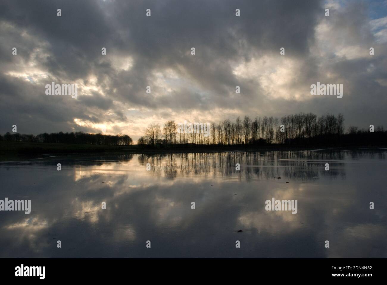 Winter scene with a line of trees along the edge of a frozen lake with light and clouds streaming from behind the trees and reflected in the ice Stock Photo