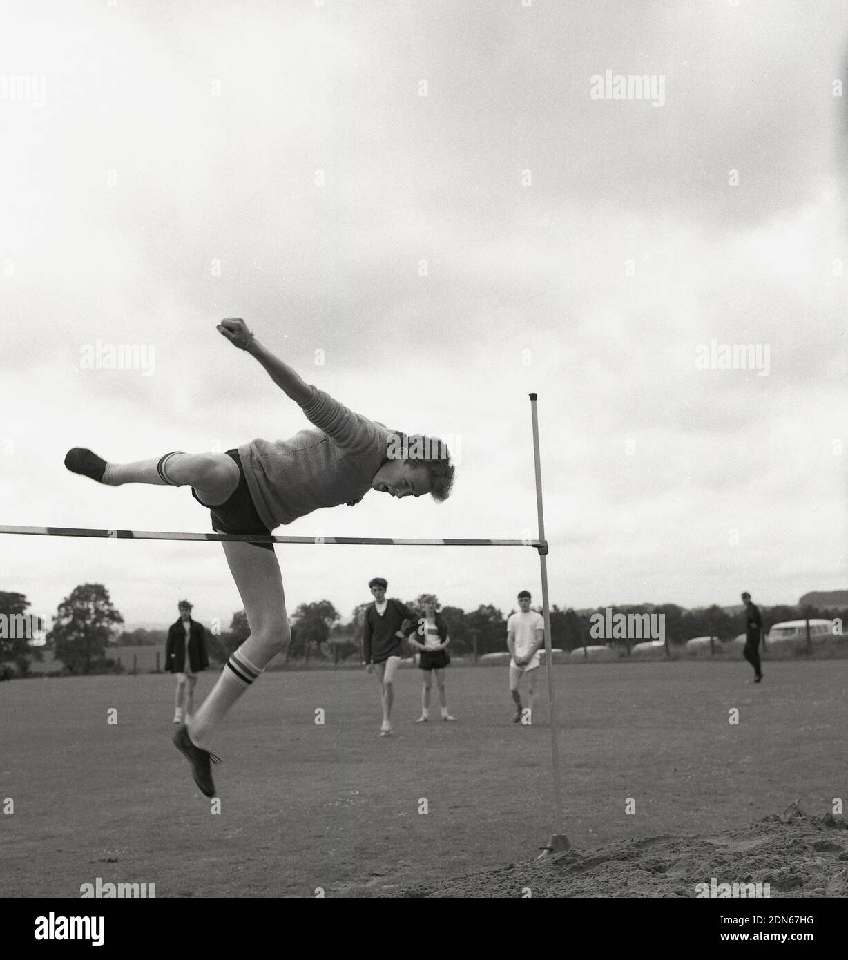 1960s, historical, outside, a young man doing the high jump at a high school sports day, using a form of straddle method, the parallel straddle, where the lead leg is kicked high in the air and the head and trunk clear the bar at the same time. The 1956 Olympic Champion, Charles Dumas - and the first jumper to clear 7ft - used this version of the straddle. The other version of the straddle is the dive method, where the head goes over the bar first. Stock Photo