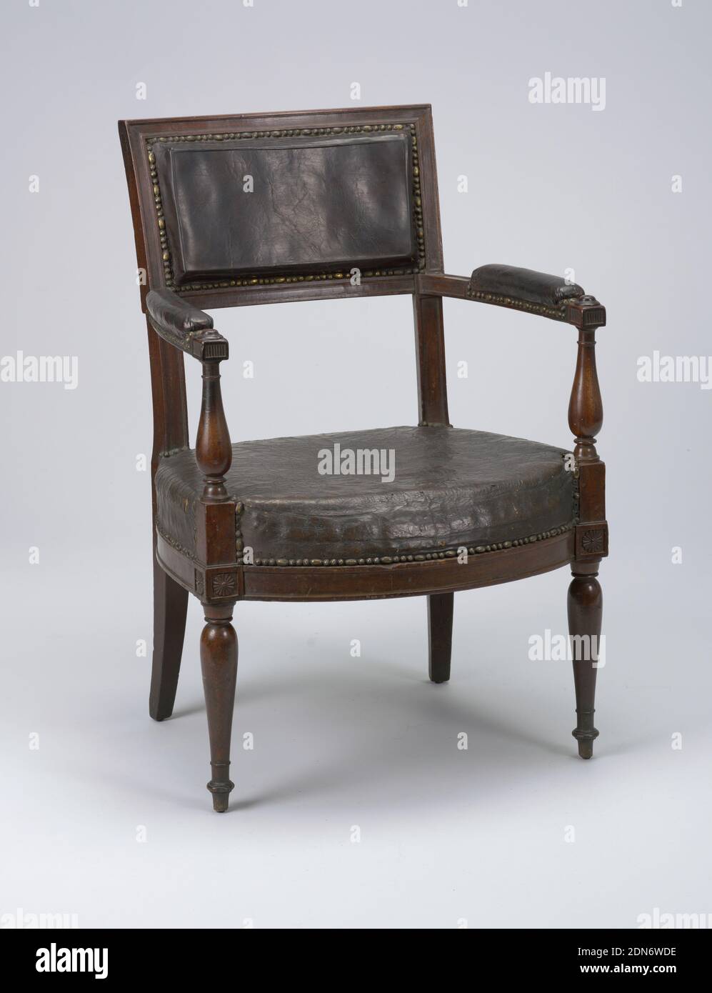 Armchair, mahogany, upholstered in nailed leather, Rectangular back. Flat padded arms curve outward and are supported by their turned stumps which are in one with the front legs, similarly turned. Bowed front and side seat rails received by rosetted reserves. Rear legs square and tapered. Upholstery is black leather nailed to frame., France, ca. 1790, furniture, Decorative Arts, Armchair Stock Photo