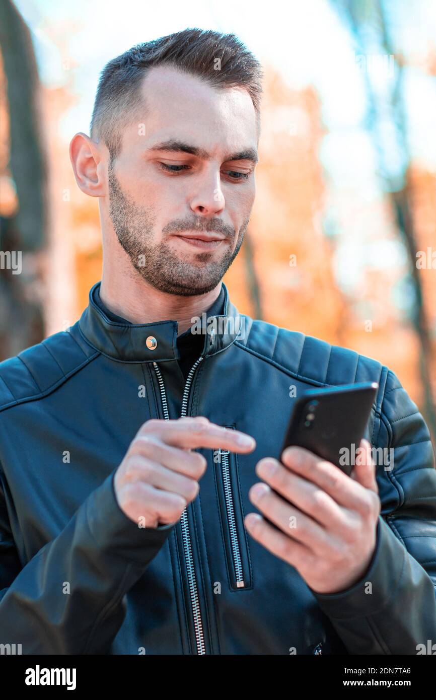 Youthful Guy Using Black Smartphone at the Beautiful Autumn Park. Handsome Young Man with Mobile Phone at Sunny Day - Medium CloseUp Shot Stock Photo