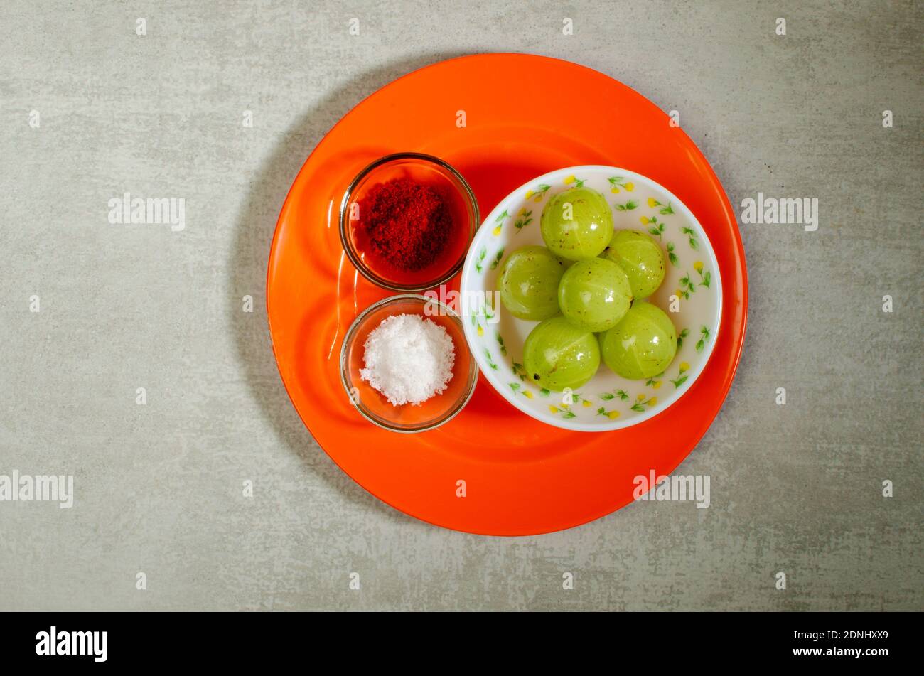 Raw Amla, Indian Gooseberry, Phyllanthus emblica. Sour in taste with Red Chilli Powder, Salt in  Orange Dish on Grey Abstract background Stock Photo