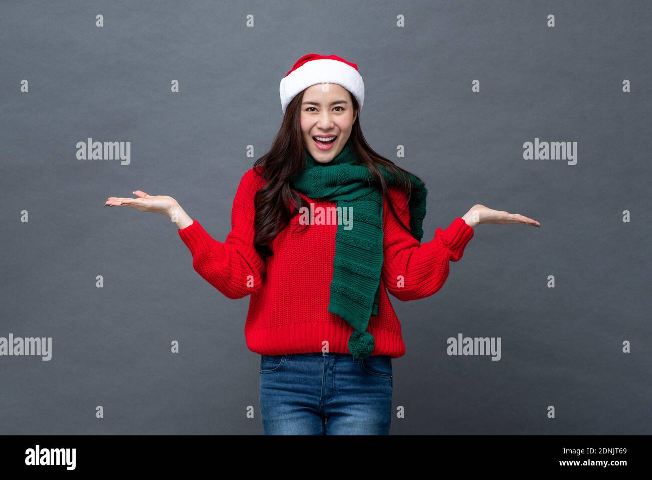 Surprised Asian woman in Christmas attire smiling and opening hands in gray isolated studio background Stock Photo
