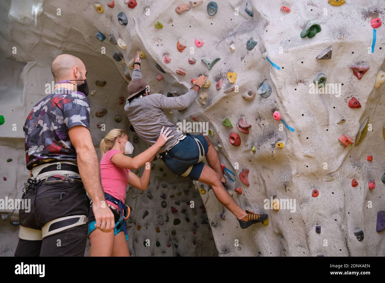Group of three young friends wearing protective face masks climbing and helping each other on artificial rock climbing wall indoors. Extreme sports in Stock Photo