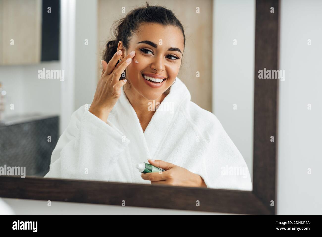 Middle east woman applying cream in bathroom mirror. Young smiling female putting moisturizer on her face. Stock Photo
