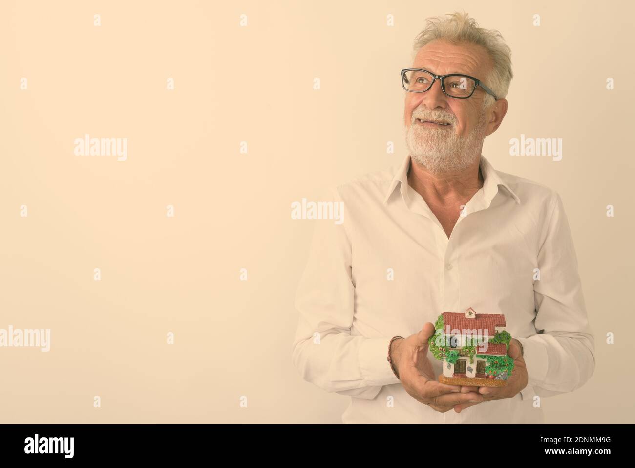 Studio shot of happy senior bearded man smiling while thinking and holding house figurine with eyeglasses against white background Stock Photo