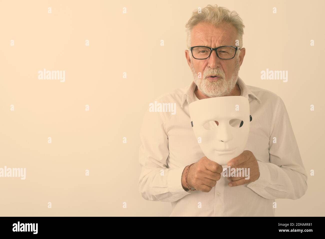 Studio shot of handsome senior bearded man holding white mask while wearing eyeglasses against white background Stock Photo