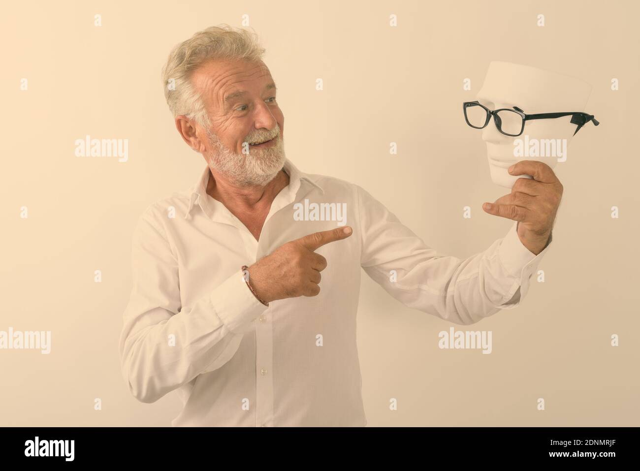 Studio shot of happy senior bearded man smiling while holding and pointing at white mask with eyeglasses against white background Stock Photo