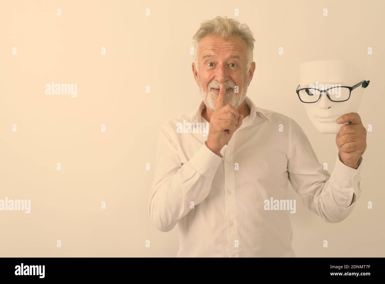Studio shot of happy senior bearded man smiling with finger on lips while holding white mask with eyeglasses against white background Stock Photo