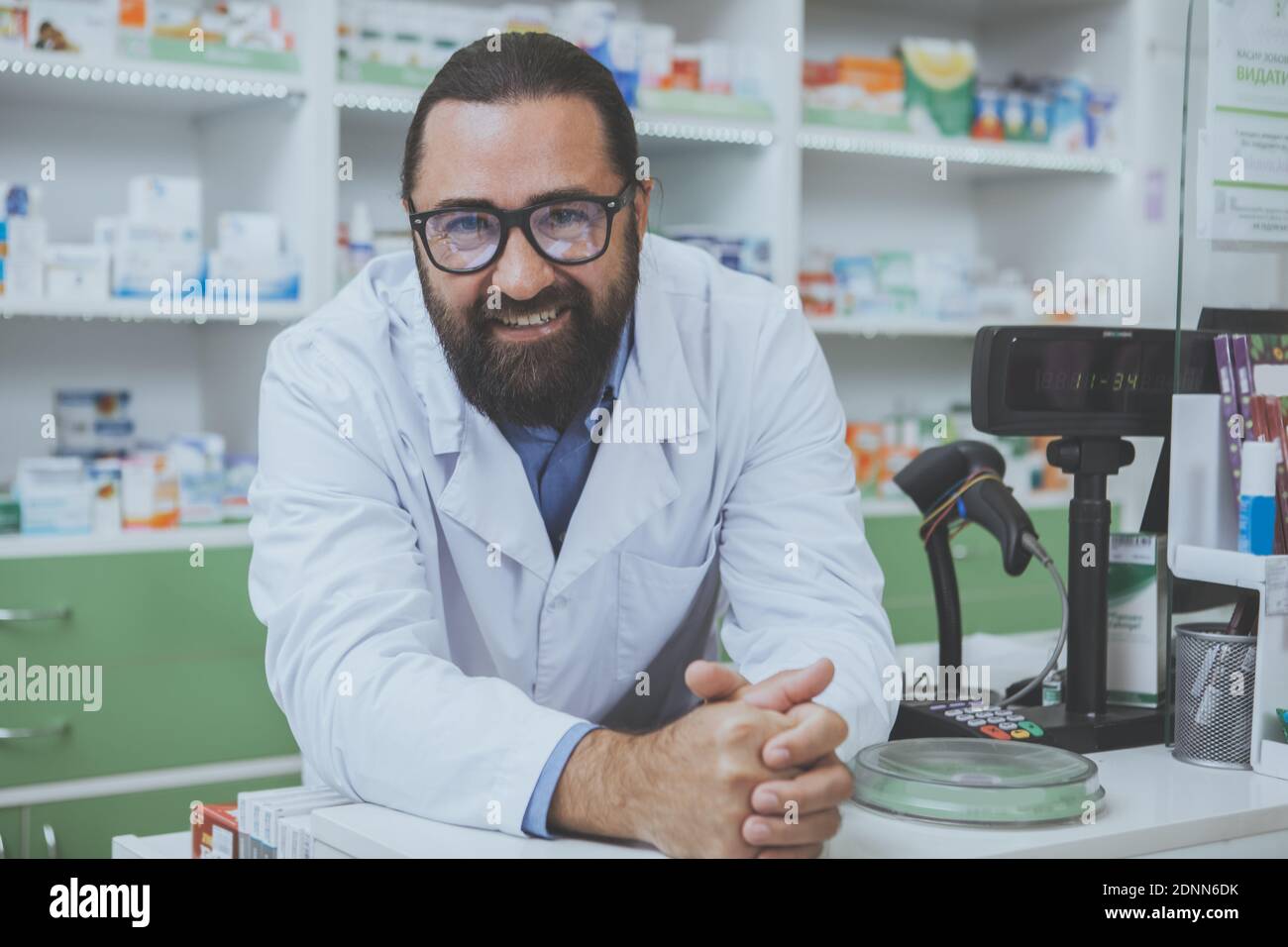 Cheerful friendly pharmacist leaning on a checkout counter at his drugstore, smiling to the camera. mature bearded druggist working at his pharmacy, c Stock Photo