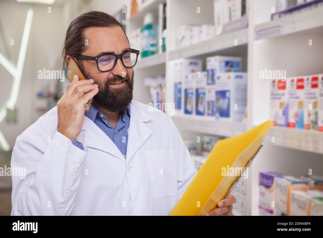 Handsome mature bearded pharmacist talking on the phone, selling medication online. CHeerful male chemist working at pharmacy, reading medical papers Stock Photo